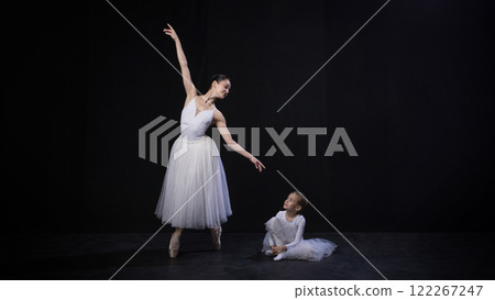 Adult ballerina raising arm gracefully, child sitting and watching attentively, both dressed in white ballet costumes, against black studio background. 122267247