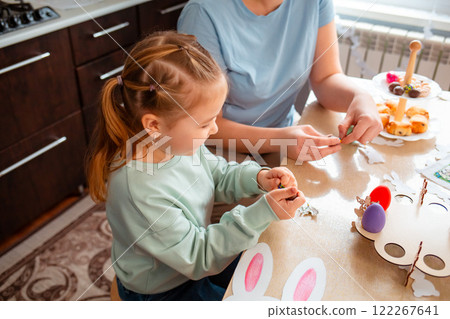 Top view of Caucasian cute little daughter and her mother are preparing at home for the Easter spring holiday. Festive table and kitchen decoration with handmade decorations 122267641