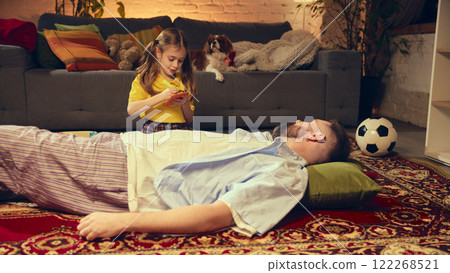 Preschool girl in yellow shirt pretends to be a doctor, writes notes on notepad while her father lies on the carpet and dog sits on couch in the background. 122268521