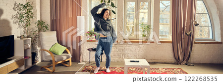 Young man in casual stylish clothes, oversized hoodie and pants, listening to music in headphone and joyfully dancing at home in living room Young man in casual stylish clothes, oversized hoodie and pants, listening to music in headphone and joyfully dancing at home in living room 122268535