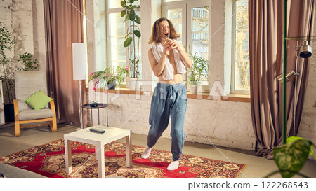 Young shirtless guy in jeans and towel on his shoulders, emotionally getting ready, signing in toothbrush and dancing in warmly lit living room Young shirtless guy in jeans and towel on his shoulders, emotionally getting ready, signing in toothbrush and dancing in warmly lit living room 122268543