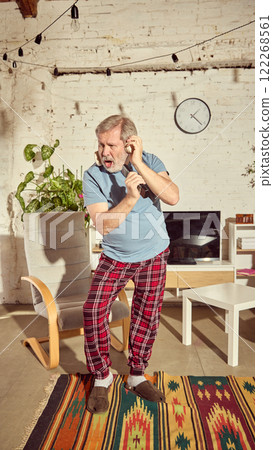 Emotional senior man in pajamas listening to music on headphone, singing in tv controller and dancing in stylish light living room at home Emotional senior man in pajamas listening to music on headphone, singing in tv controller and dancing in stylish light living room at home 122268561
