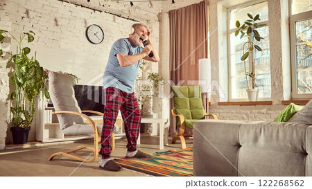 Elderly man in pajamas listening to music in headphones and emotionally singing in tv controller in warmly lit living room with plants, chair and large tv Elderly man in pajamas listening to music in headphones and emotionally singing in tv controller in warmly lit living room with plants, chair and large tv 122268562