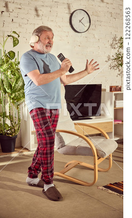 Elderly man in pajamas listening to music in headphones and singing in tv controller in warmly lit living room with plants, chair and large tv Elderly man in pajamas listening to music in headphones and singing in tv controller in warmly lit living room with plants, chair and large tv 122268563