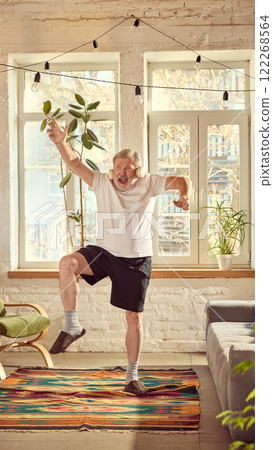 Emotional and energetic elderly man in home wear listening to music in headphones and emotionally dancing in sunny living room at home Emotional and energetic elderly man in home wear listening to music in headphones and emotionally dancing in sunny living room at home 122268564
