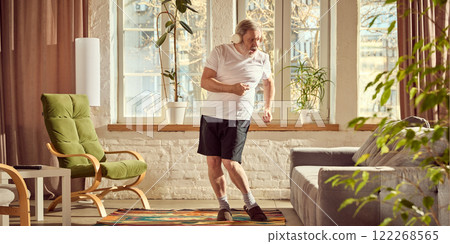 Elderly man in home wear listening to music in headphones and emotionally dancing in cozy, warmly lit living room at home with potted plants and grey couch Elderly man in home wear listening to music in headphones and emotionally dancing in cozy, warmly lit living room at home with potted plants and grey couch 122268565