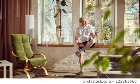 Elderly man in home wear listening to music in headphones , sitting on windowsill and playing imaginative guitar in cozy, warmly lit living room at home Elderly man in home wear listening to music in headphones , sitting on windowsill and playing imaginative guitar in cozy, warmly lit living room at home 122268566