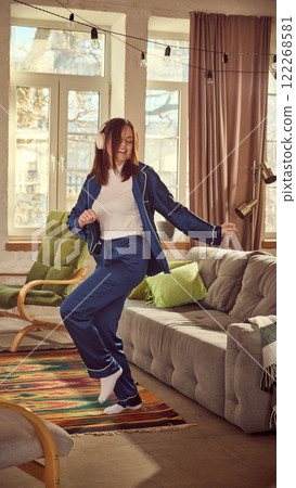 Happy, smiling young woman in blue pajamas and white top, wearing headphones, listening to music and dancing in cozy living room Happy, smiling young woman in blue pajamas and white top, wearing headphones, listening to music and dancing in cozy living room 122268581