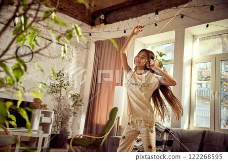 Enjoyment of weekends. Happy, smiling teen girl with headphones, listening to music and joyfully dancing in modern living room with cozy interior 122268595