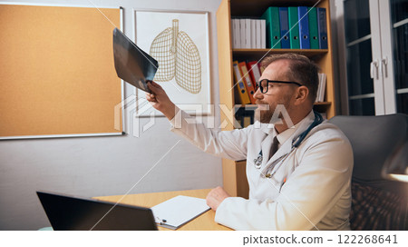 Bearded man in medical coat, glasses and stethoscope, doctor sitting at table in his office and looking at x-ray scan. Examination 122268641