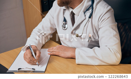 Cropped image of bearded man , doctor in white coat and stethoscope sitting at table and prescribing treatment for patient, taking notes Cropped image of bearded man , doctor in white coat and stethoscope sitting at table and prescribing treatment for patient, taking notes 122268643