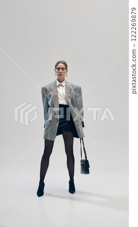 Confident businesswoman in oversized grey blazer, white shirt, leather skirt, glasses and bag, posing against light grey studio background 122269879