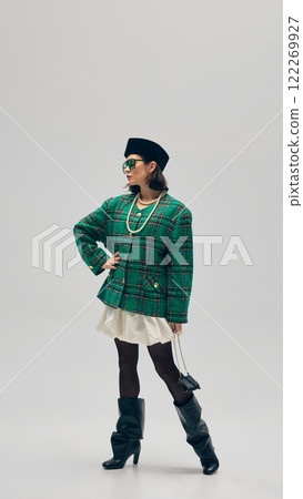 Elegant young woman wearing plaid green jacket, pearl necklace, white skirt, heeled boots, beret and sunglasses against minimalist studio background Elegant young woman wearing plaid green jacket, pearl necklace, white skirt, heeled boots, beret and sunglasses against minimalist studio background 122269927