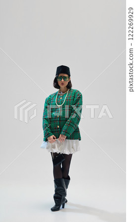 Full-length of elegant young woman with dark short hair, in green plaid blazer, white skirt, beret, heeled boots and sunglasses against studio background Full-length of elegant young woman with dark short hair, in green plaid blazer, white skirt, beret, heeled boots and sunglasses against studio background 122269929