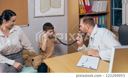 Playful and caring moment between child and doctor. Boy sitting with his mother in medical office during consultation and listening to bearded man with stethoscope Playful and caring moment between child and doctor. Boy sitting with his mother in medical office during consultation and listening to bearded man with stethoscope 122269953
