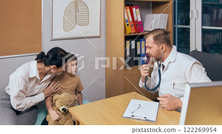 Male pediatrician with beard using stethoscope while examining preschool boy against organized office background. Child came to doctor with his mother Male pediatrician with beard using stethoscope while examining preschool boy against organized office background. Child came to doctor with his mother 122269954
