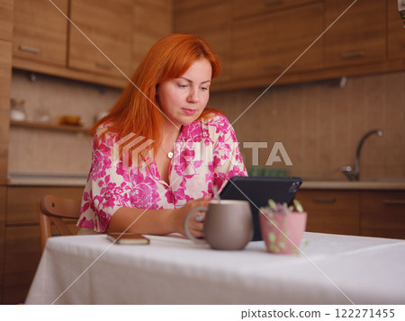 young woman working on tablet at home kitchen. A real female freelancer uses computer to work remotely from home with cup of coffee. 122271455