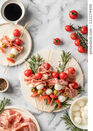 Skewers with mozzarella, ham, olives, peppers, tomatoes. Festive canapes or antipasti on white tray for party, top view 122274409