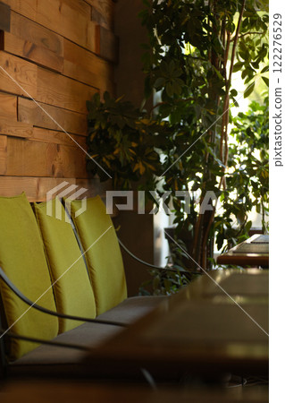 Cozy interior of a cafe with a wooden wall, green cushioned chairs, and lush indoor plants creating a relaxing and inviting atmosphere. 122276529