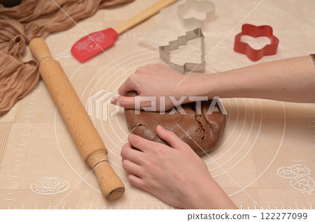 Christmas homemade baking, woman kneading brown cocoa dough with her hands for baking gingerbread, shaped cutter, silicone spatula and rolling pin on the table 122277099
