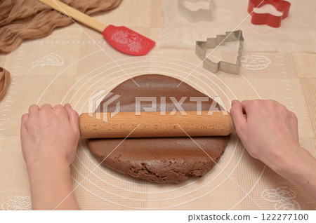 Christmas homemade baking, woman roll out brown cocoa dough with rolling pin for baking gingerbread, shaped cutter and silicone spatula on the table 122277100