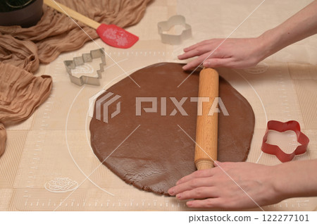 Christmas homemade baking, woman roll out brown cocoa dough with rolling pin for baking gingerbread, shaped cutter and silicone spatula on the table 122277101