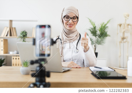 Middle Eastern female doctor wearing hijab recording online medical lesson, providing advice on smartphone. Young professional in modern office setting, promoting healthcare through digital platform. Middle Eastern female doctor wearing hijab recording online medical lesson, providing advice on smartphone. Young professional in modern office setting, promoting healthcare through digital platform. 122282383