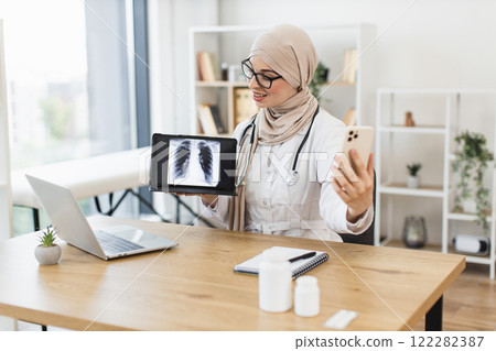 Muslim female doctor performs video call with patient, displaying lung x-ray on tablet. Middle-aged professional wearing stethoscope in medical office setting. Muslim female doctor performs video call with patient, displaying lung x-ray on tablet. Middle-aged professional wearing stethoscope in medical office setting. 122282387
