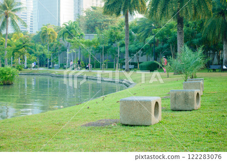 serene park scene featuring stone benches by tranquil lake, surrounded by lush greenery and palm trees. People enjoy leisure activities in background, creating peaceful atmosphere 122283076