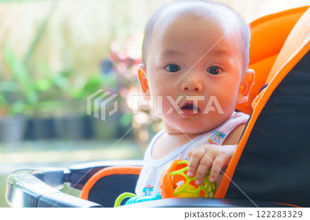 curious baby boy sitting in stroller, holding colorful toy, surrounded by greenery. His expression shows wonder and joy in bright outdoor setting 122283329