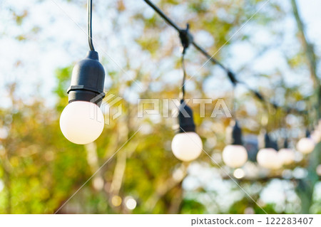 Bright outdoor string lights hanging in garden, creating warm atmosphere. blurred background features trees and greenery, enhancing cozy ambiance Bright outdoor string lights hanging in garden, creating warm atmosphere. blurred background features trees and greenery, enhancing cozy ambiance 122283407