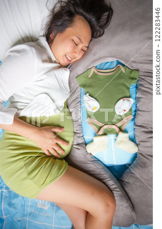 pregnant woman lying on bed, smiling joyfully while holding her belly, with green baby onesie and knitted baby booties placed beside her, symbolizing anticipation and love pregnant woman lying on bed, smiling joyfully while holding her belly, with green baby onesie and knitted baby booties placed beside her, symbolizing anticipation and love 122283446
