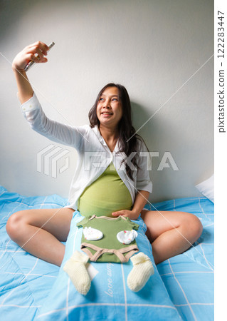 pregnant woman joyfully takes selfie while sitting on bed, with baby clothes and accessories placed on her belly, symbolizing excitement and preparation for her baby pregnant woman joyfully takes selfie while sitting on bed, with baby clothes and accessories placed on her belly, symbolizing excitement and preparation for her baby 122283447