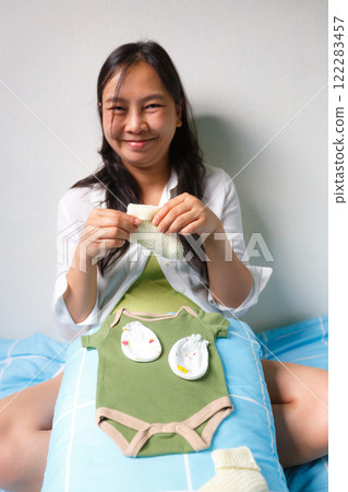 smiling woman sits on bed holding knitted baby sock, with baby clothes and accessories placed on her lap, creating warm and joyful atmosphere smiling woman sits on bed holding knitted baby sock, with baby clothes and accessories placed on her lap, creating warm and joyful atmosphere 122283457