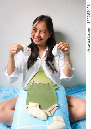smiling pregnant woman sits on bed holding baby socks, with green baby onesie and yellow socks placed on pillow in front of her, expressing joy and anticipation 122283458