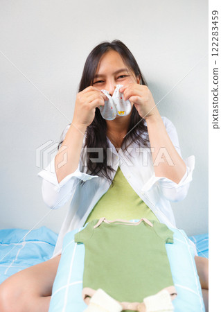 pregnant woman sitting on bed, smiling joyfully while holding baby socks, with green baby onesie placed on her lap, expressing excitement and anticipation for her baby 122283459