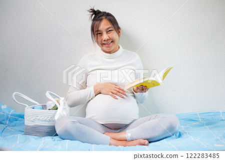 pregnant woman reading book while sitting on bed, surrounded by baby items. She looks happy and relaxed, enjoying her time preparing for motherhood pregnant woman reading book while sitting on bed, surrounded by baby items. She looks happy and relaxed, enjoying her time preparing for motherhood 122283485