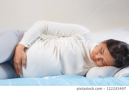 woman lying on bed, holding her belly, showing signs of discomfort. She is wearing long sleeve top and leggings, with calm expression woman lying on bed, holding her belly, showing signs of discomfort. She is wearing long sleeve top and leggings, with calm expression 122283497