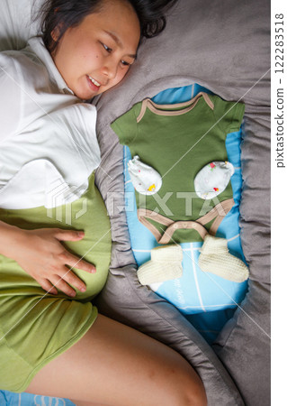 pregnant woman lies on bed, smiling while looking at baby clothes, including green onesie, mittens, and socks, symbolizing anticipation and joy for upcoming baby 122283518