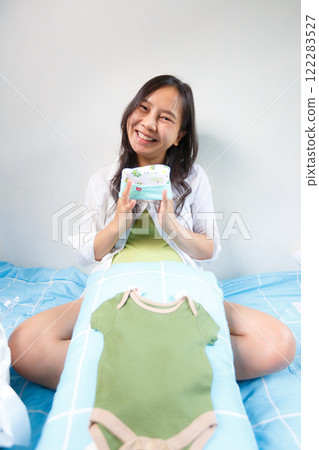 smiling woman sitting on bed holding pack of baby diapers, with green baby onesie placed on pillow in front of her, creating joyful and nurturing atmosphere 122283527
