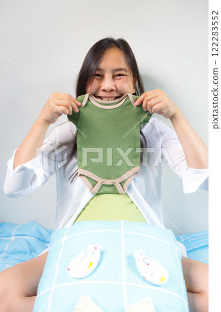 smiling pregnant woman holding green baby onesie while sitting on bed with baby shoes and blanket, expressing joy and excitement for her upcoming baby smiling pregnant woman holding green baby onesie while sitting on bed with baby shoes and blanket, expressing joy and excitement for her upcoming baby 122283552