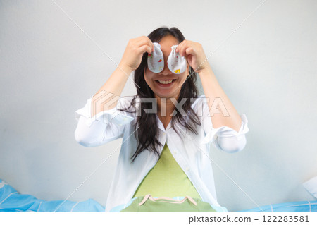 pregnant woman sitting on bed playfully holding baby socks over her eyes, expressing joy and excitement for her upcoming baby 122283581