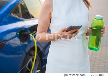 Woman in casual wear checks her phone and holds a water bottle while charging an EV Woman in casual wear checks her phone and holds a water bottle while charging an EV 122283800