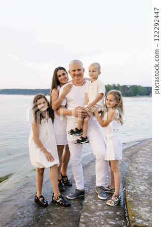 Happy family of five is standing on stone staircase by a body of water. The children are wearing white dresses 122292447