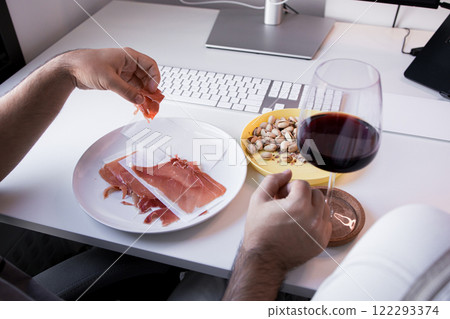 Man enjoys prosciutto, pistachios, and red wine at office desk 122293374