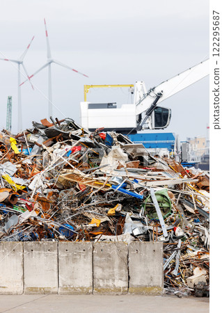Crane at recycling facility loading large scrap metal pile with wind turbines in background. Industrial area scene waste management and resource reuse concept 122295687