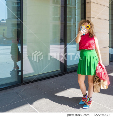 a beautiful young woman in bright clothes with a phone and bags takes selfies or takes pictures of the city on a walk a beautiful young woman in bright clothes with a phone and bags takes selfies or takes pictures of the city on a walk 122296672