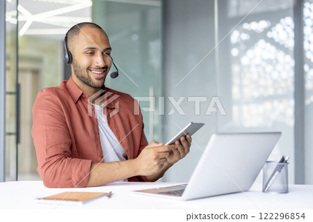 Man with headset working inside office, using tablet computer and laptop. Support service worker at workplace, consulting and helping clients, online remotely. 122296854