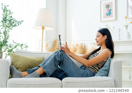 Side view and wide shot beautiful Asian woman lean on sofa also enjoy to use tablet in living room with day light. 122298540