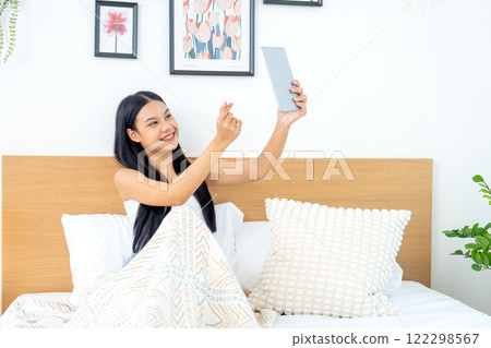 Beautiful Asian woman lean on bed with bend her knees also hold tablet and show sign of love use hands with relax and happiness in bedroom. 122298567